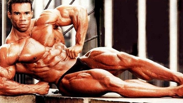 Bodybuilder Kevin Levrone reclining, shirtless, abs visible and contacting his muscles.