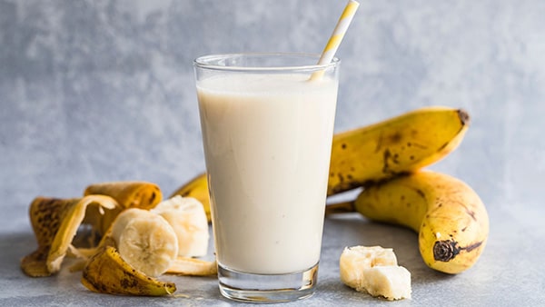 A banana smoothie in a tall glass with a straw, and bananas placed behind it.