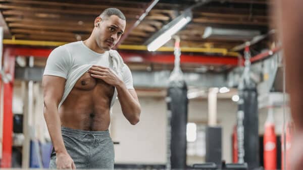 Un homme se regarde dans le miroir d'une salle de sport en relevant son t-shirt, ventre et pectoraux apparents.