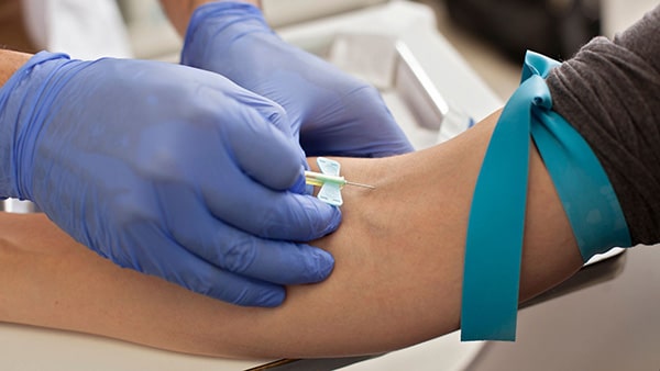 A doctor wearing blue gloves pricks a vein in a person's right arm for a blood test.