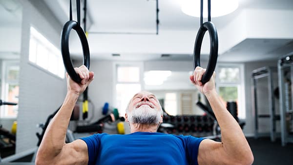 A man in his fifties, wearing a blue T-shirt, does an upper-body exercise with rings in a gym.