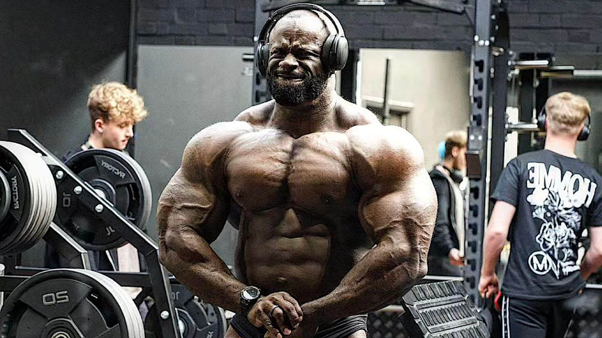 Professional bodybuilder Samson Dauda, shirtless in a weight room, posing with headphones on.