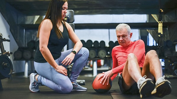 In a weight room, a man over 50 does abdominal exercises, seated on a fitness mat, assisted by a sports coach.