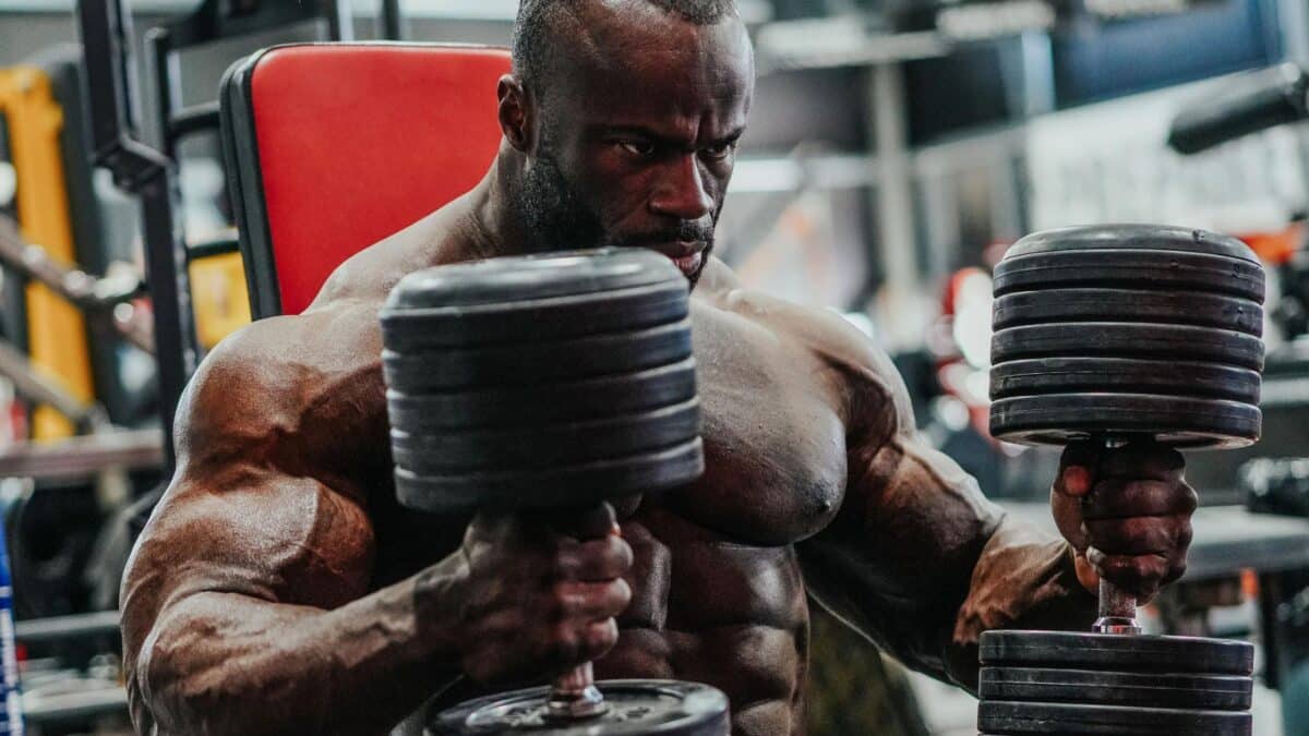 Pro bodybuilder Samson Dauda in a gym, sitting on a weight bench with two dumbbells in his hands.