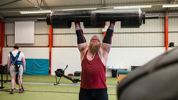 Dans une salle de sport, un strongman en débardeur fait du log press en soulevant au-dessus de sa tête, bras tendus, un objet lourd.