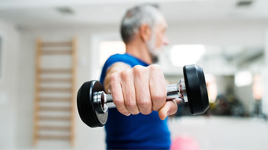 A man over 50 exercises in a gym, doing lateral raises with dumbbells.