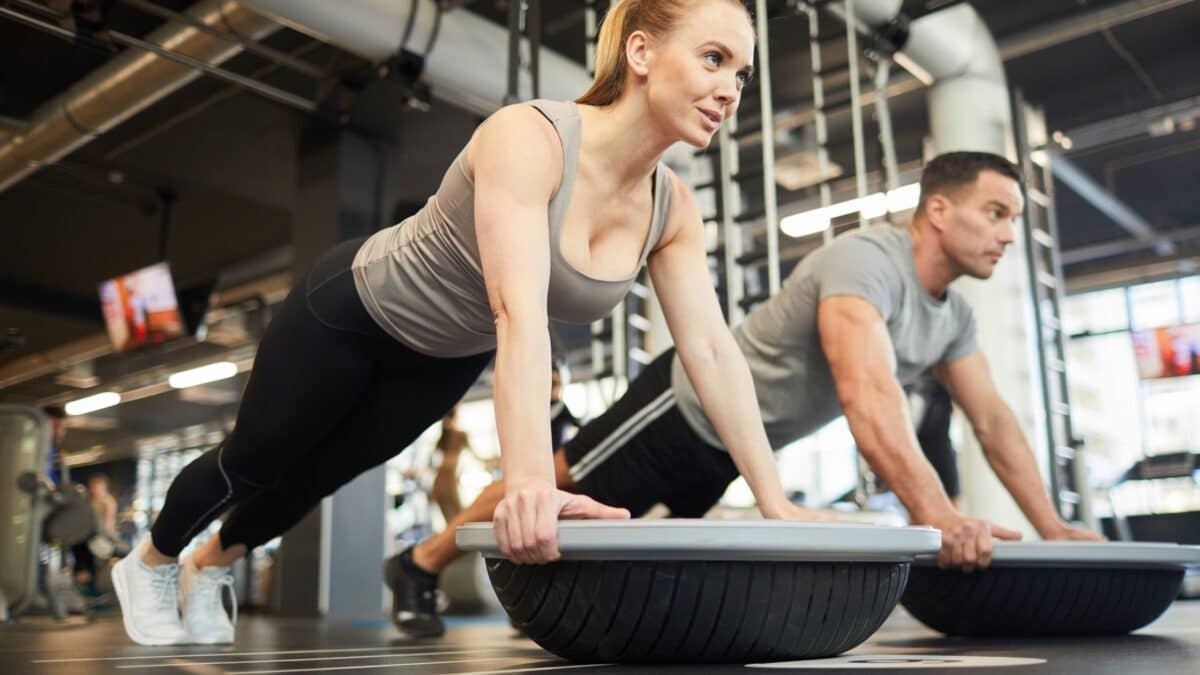 Dans une salle de musculation, un homme et une femme en tenues de sport font de la proprioception pour le haut du corps, en position de planche bras tendus sur un BOSU.