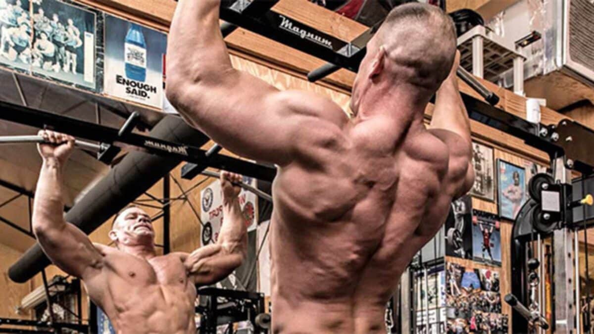 John Cena shirtless, doing pull-ups in a weight room.