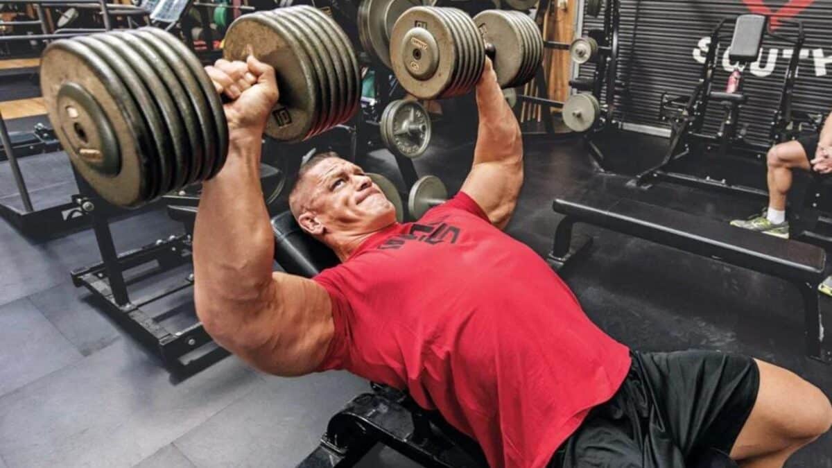 Professional wrestler John Cena doing a dumbbell bench press in a weight room.