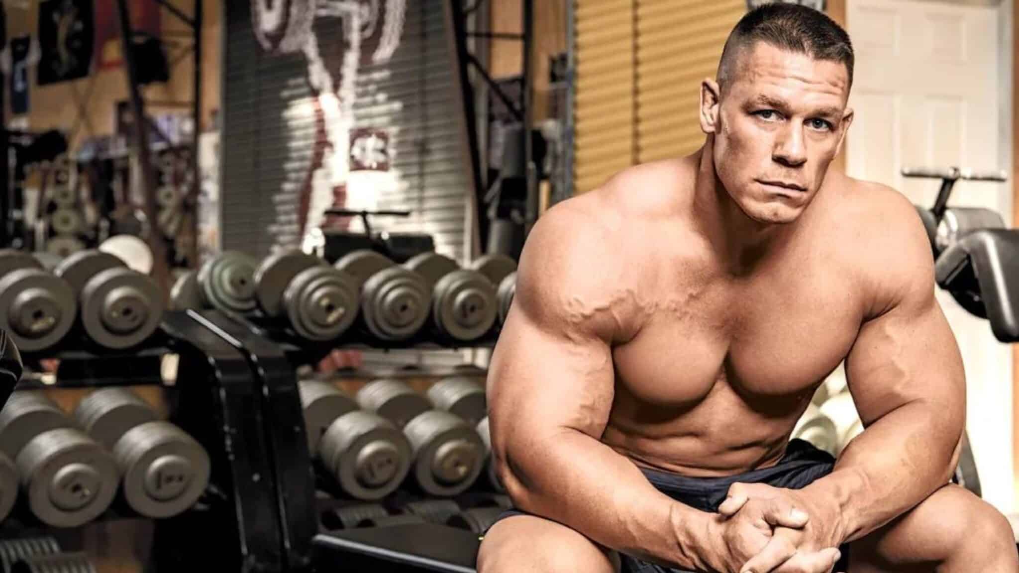 Shirtless athlete John Cena sits on a weight bench in a gym.