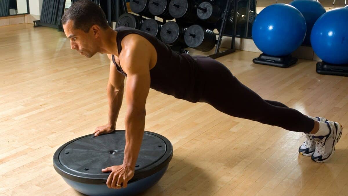 A man in a tank top does push-ups on a BOSU in a weight room.