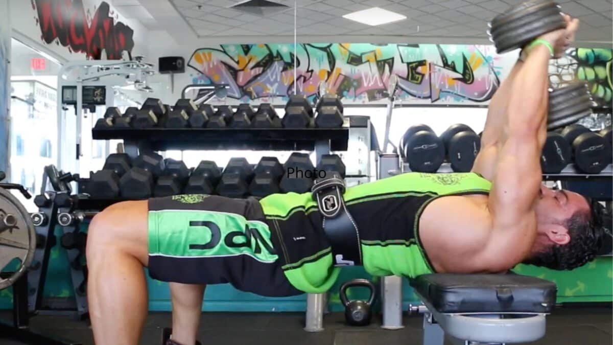 Fitness trainer Julien Quaglierini pulls over in a weight room, with his back stretched out on a weight bench and a dumbbell in his hands.