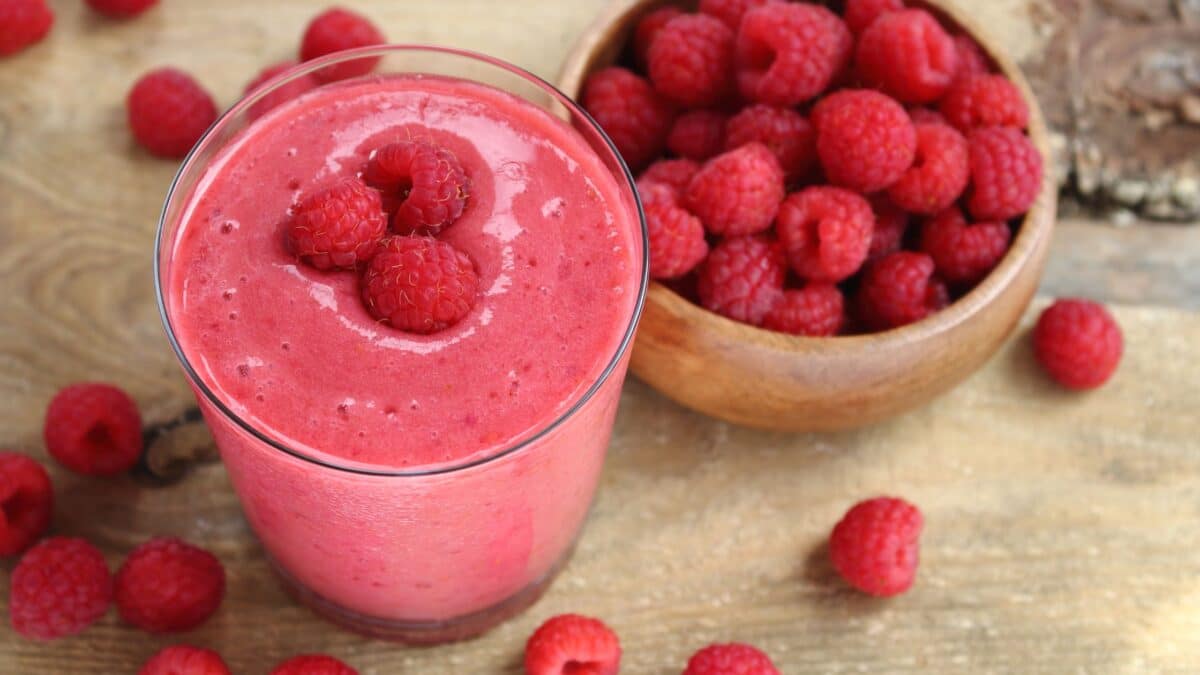 A glass containing a pink protein drink, with a bowl of raspberries alongside.