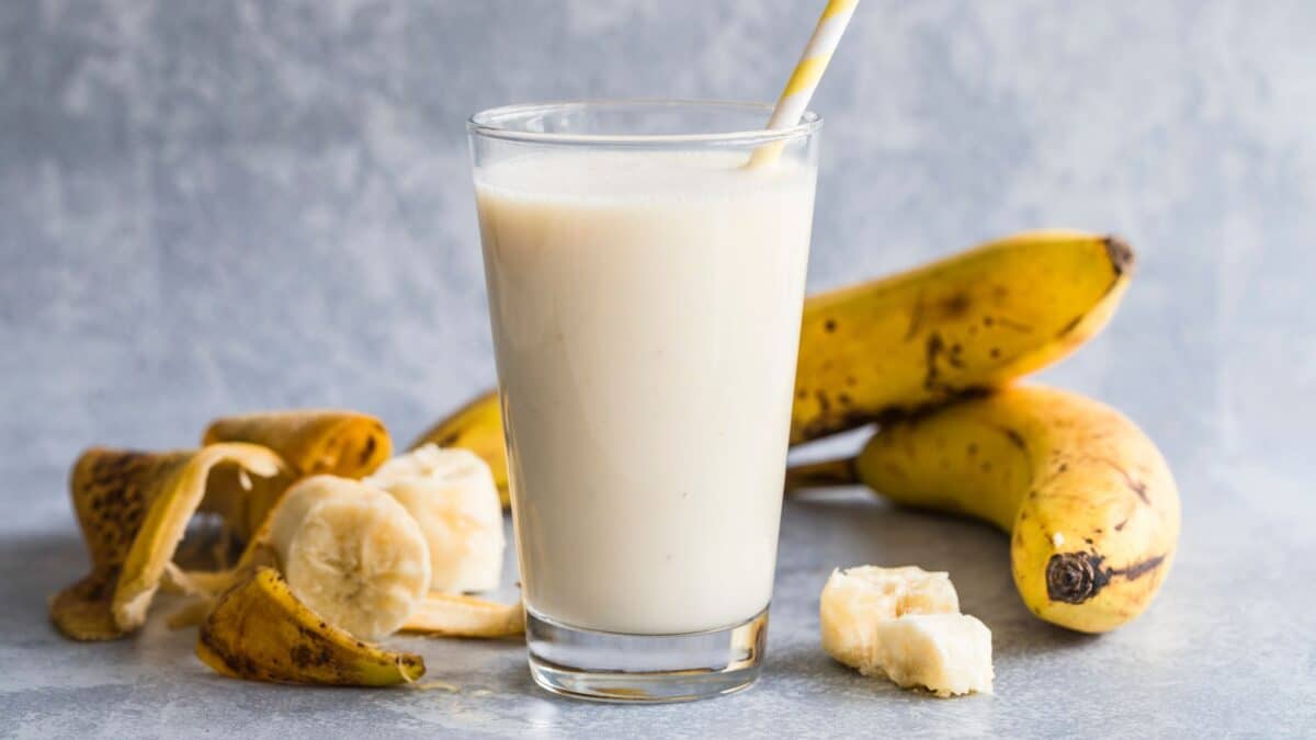 A clear glass with a straw, in front of several bananas, in which there's a vanilla protein drink.