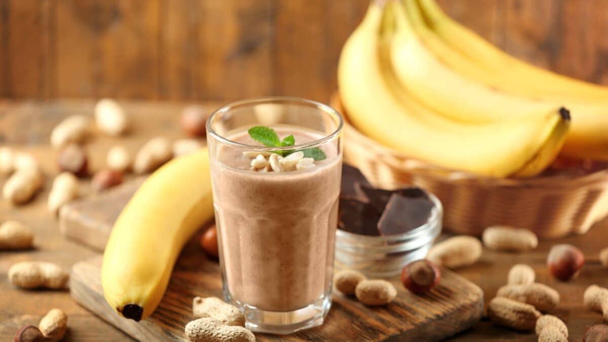 A glass containing a brown protein drink, next to which are bananas, hazelnuts and a bowl of dark chocolate.