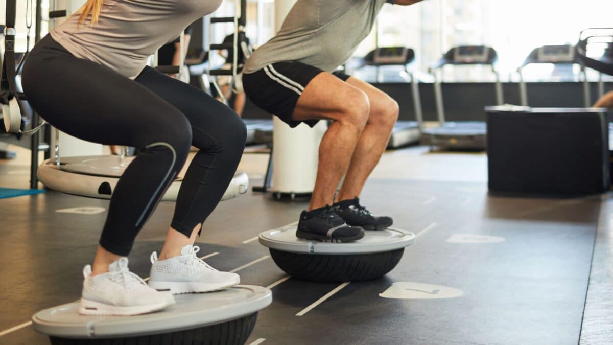 In a gym, a man and a woman do squats on a BOSU to improve their proprioception.