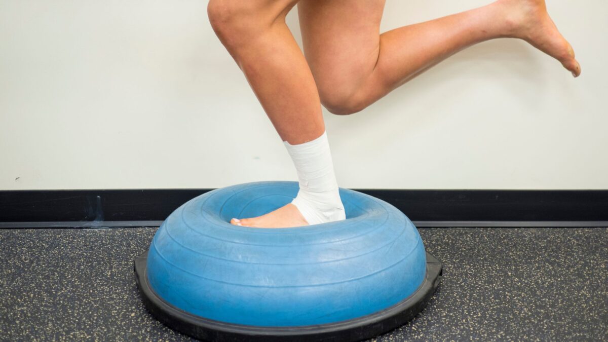 Un homme avec la cheville gauche bandée fait un exercice d'équilibre avec sa jambe blessée posée sur la face molle d'un BOSU.