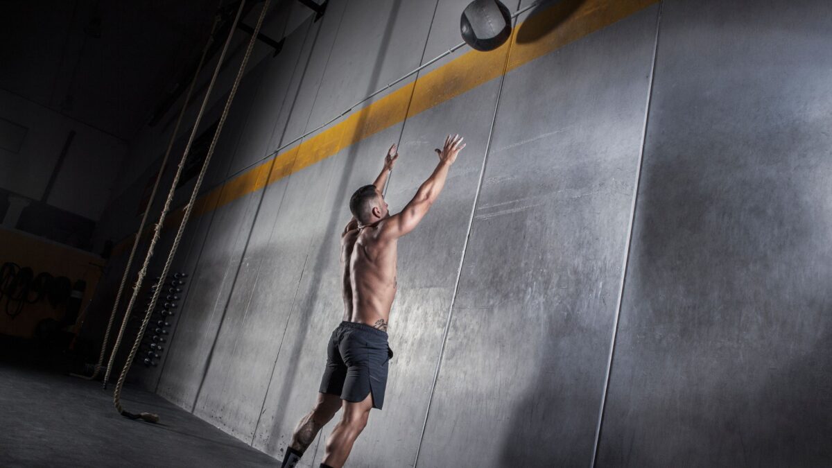 Un homme torse nu musclé fait l'exercice du wall ball avec un médecine ball contre un mur.