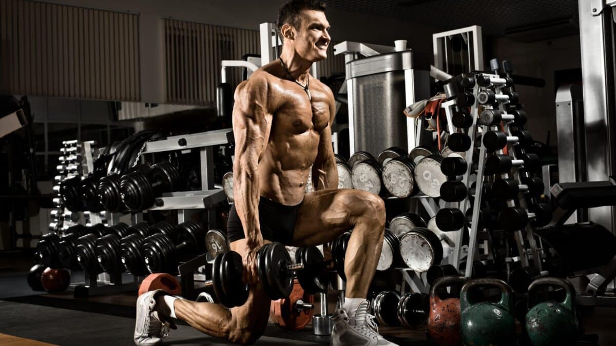 A shirtless man does lunges with dumbbells in his hands in a weight room.