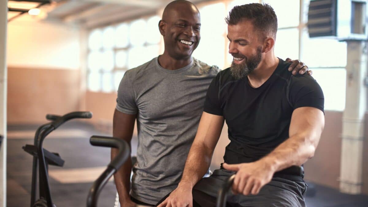 Two men smile as they chat in a fitness room, one standing, the other sitting on an elliptical.