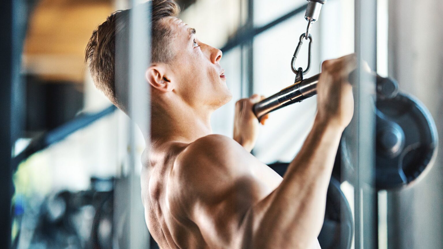 Un homme torse nu fait un exercice de dos à la poulie dans une salle de musculation : du tirage vertical.