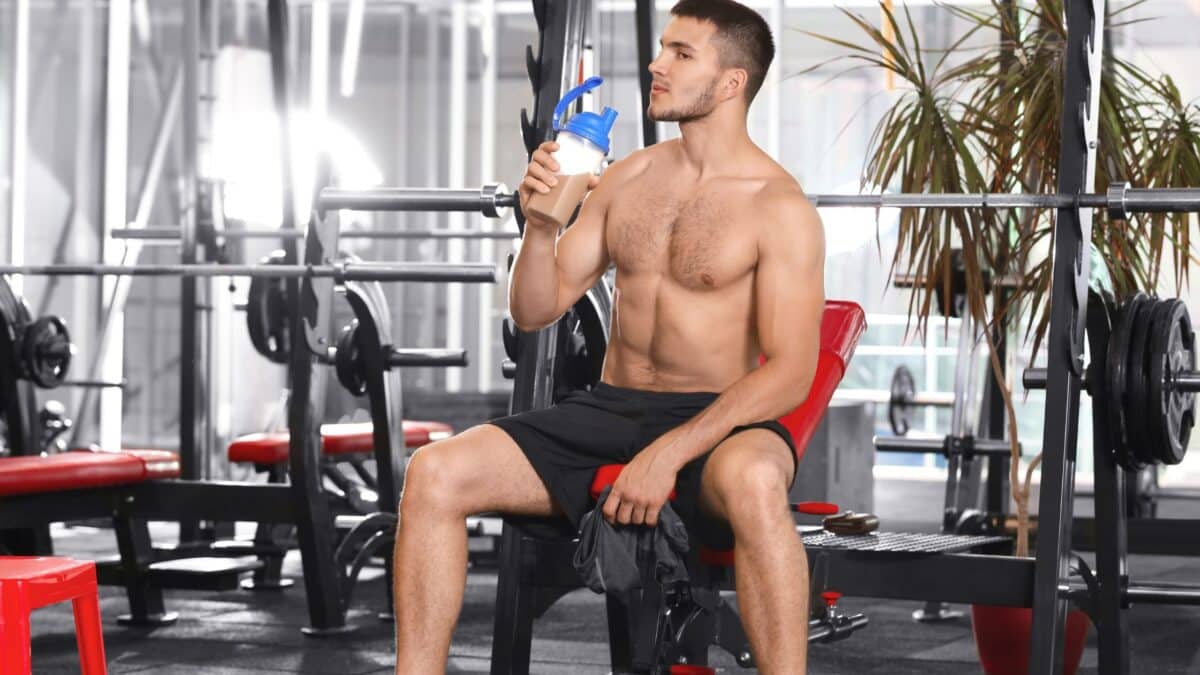 A shirtless bodybuilder sitting on a bench press in a gym drinks from a shaker.