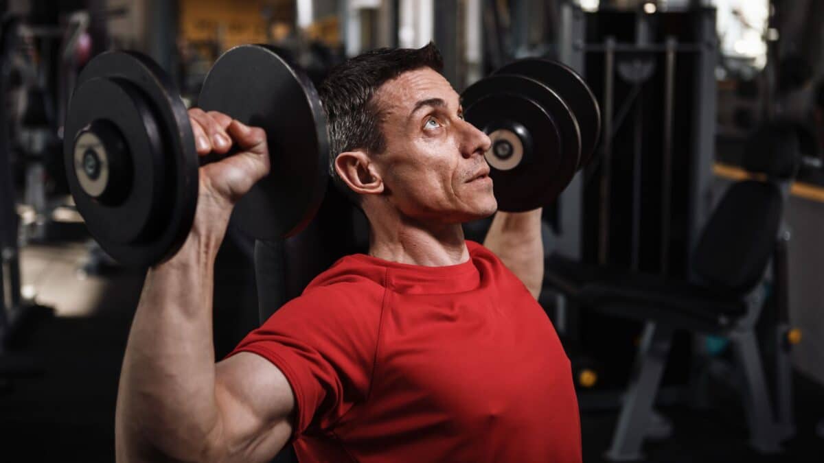 A man in his 40s performs a military bench press with dumbbells in a weight room.