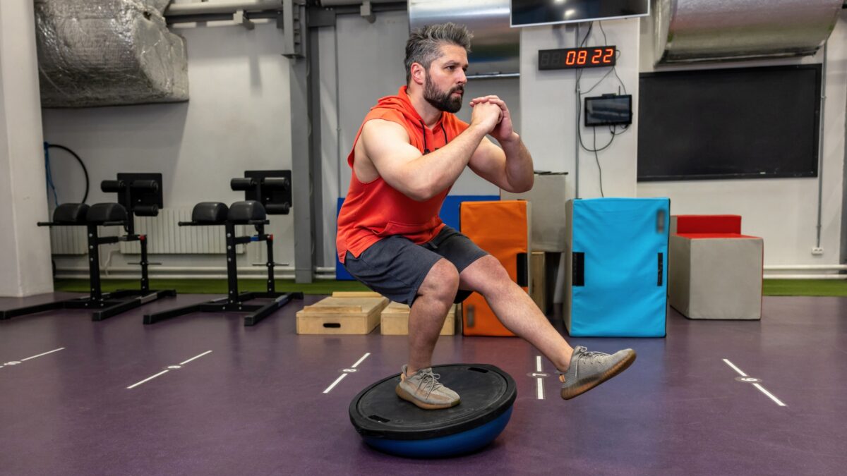 Dans une salle de sport, un homme en short fait un exercice de proprioception, debout sur une jambe sur un BOSU.