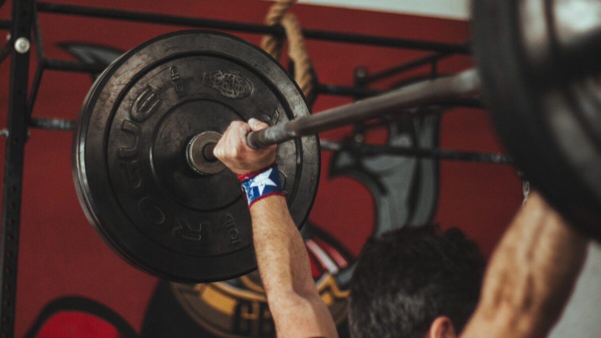 Un pratiquant de crossfit fait l'exercice du push press avec une barre de musculation chargée, les bras tendus au-dessus de la tête.