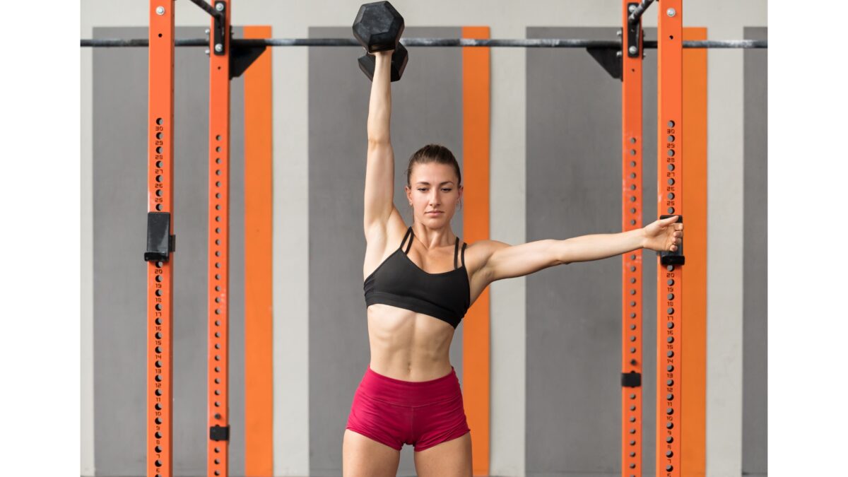 Une femme en tenue de sport fait l'exercice du push press avec un haltère dans la main droite, bras tendu au-dessus de sa tête.