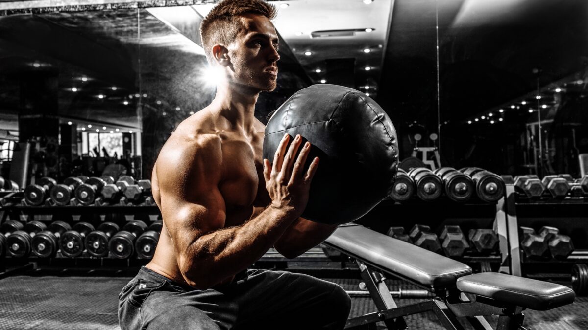 Dans une salle de musculation, un homme torse nu et musclé fait l'exercice du wall ball.