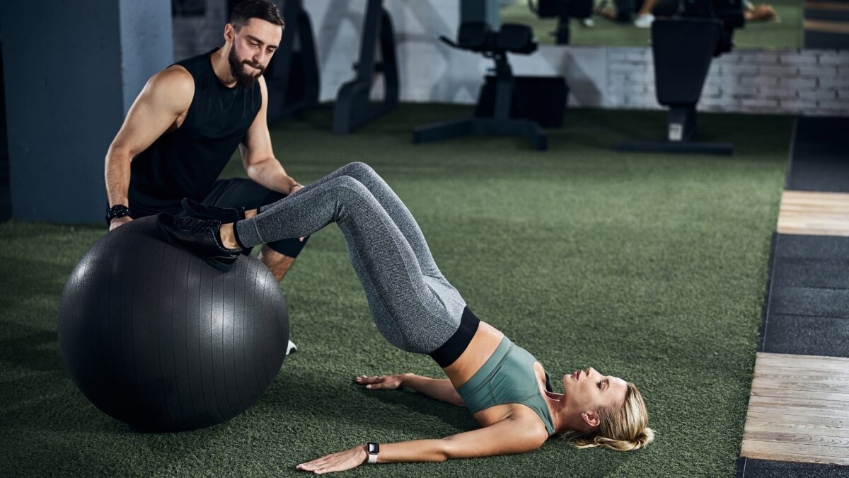 Une femme fait du leg curl sur ballon suisse avec un coach de sport dans une salle de fitness, un bon exercice pour les ischios.